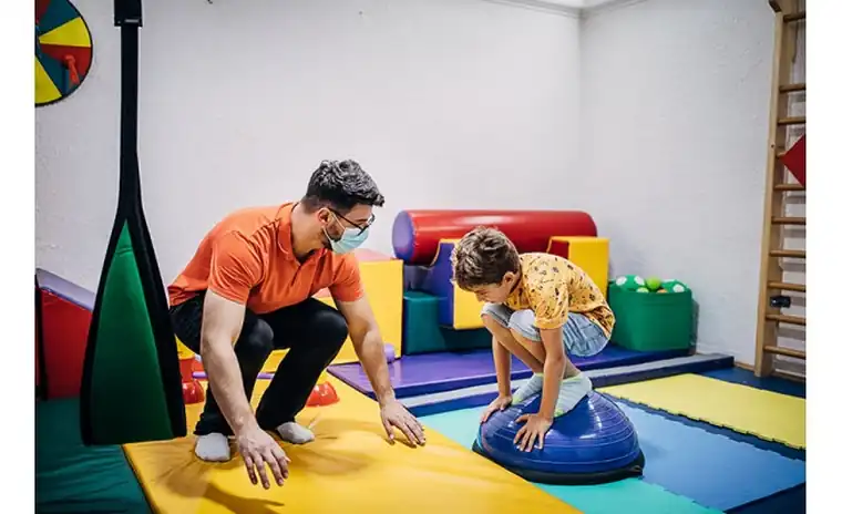 Kinésithérapeute guidant un enfant lors d’un exercice d’équilibre sur ballon dans une salle de rééducation pédiatrique